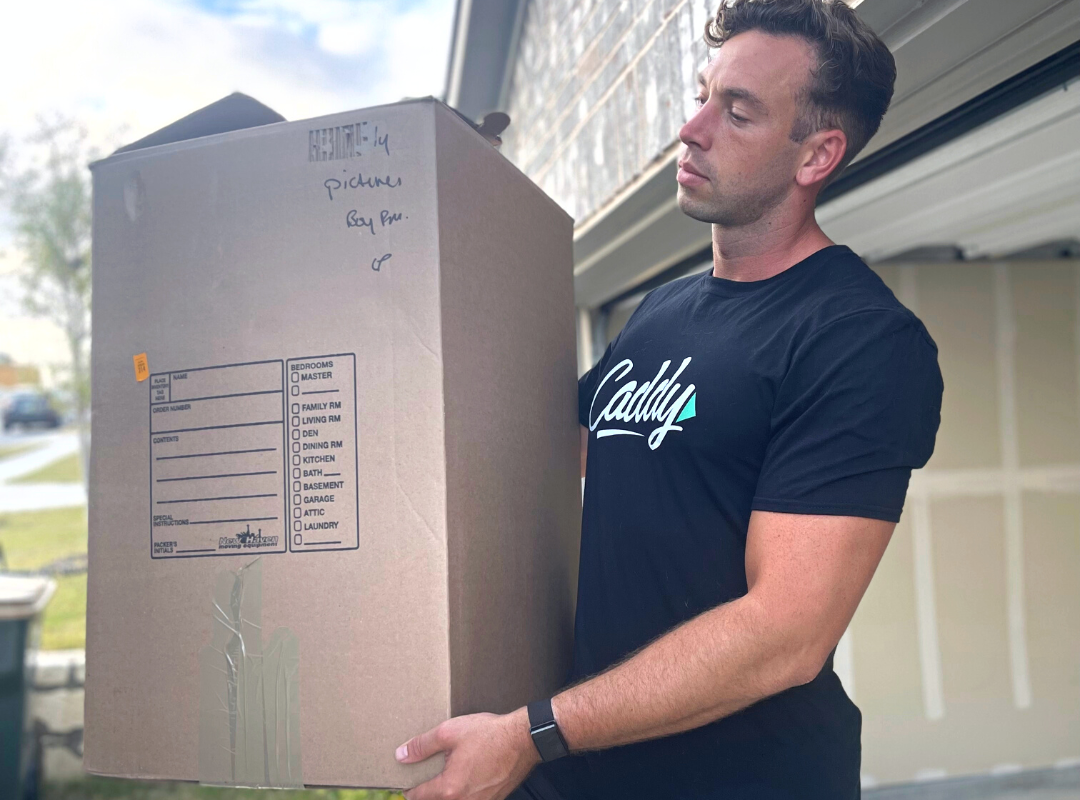 Caddy mover, Chris, lifting a heavy moving box into a rental moving truck outside of a suburban home