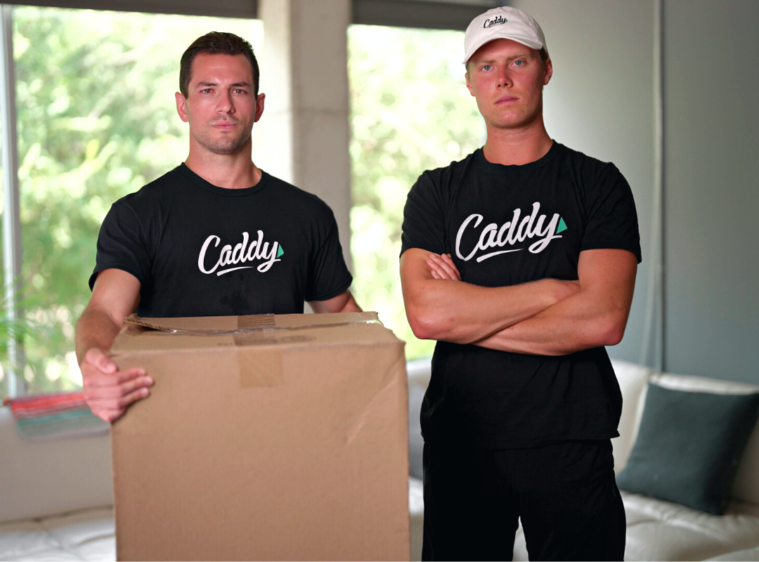 Zach and Ben, Movers from Caddy, looking strong, willing, and ready to move. They are staring at the camera holding a moving box with serious faces.