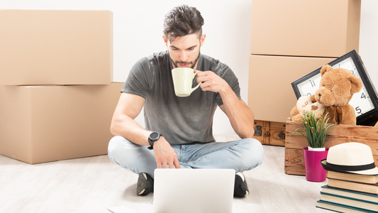 A man moving alone with his first apartment, looking at his laptop while sipping coffee.