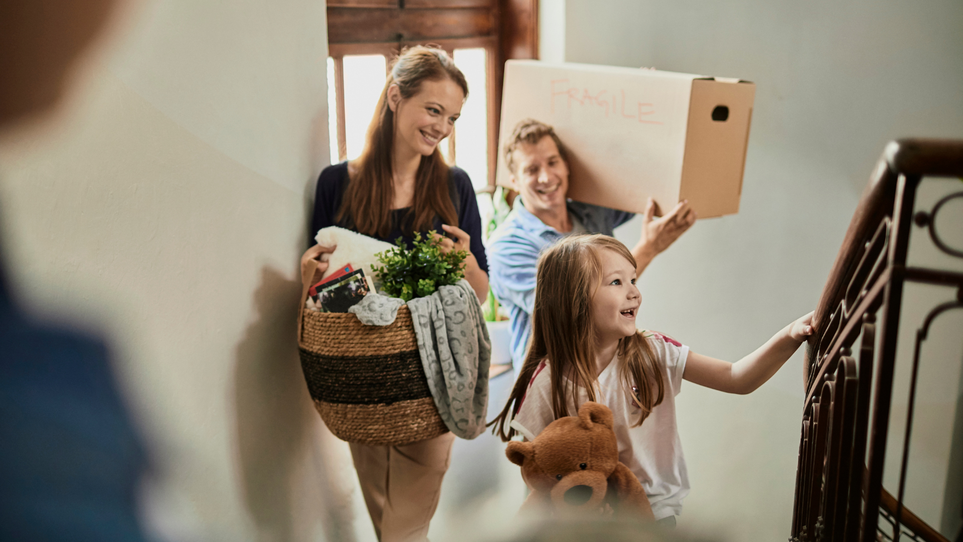 Family of three just arrive in their new home. Little carrying her teddy followed by her mom carrying a basket load of stuff and dad carrying a box with fragile items inside