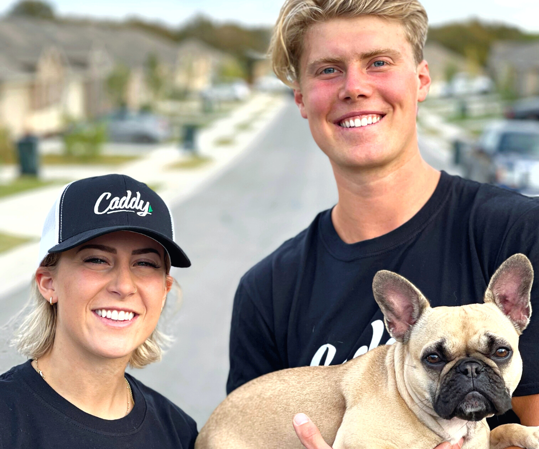 Caddy movers, Zach and Emily, holding the Caddy mascot dog, Kiki, smiling outside of a family suburb